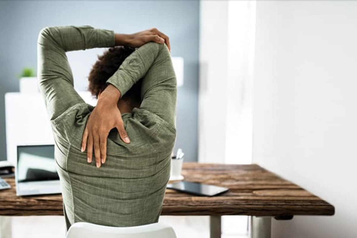 Woman in green long sleeve shirt sitting at home office desk with back to camera performing stretches for scapular dyskinesis