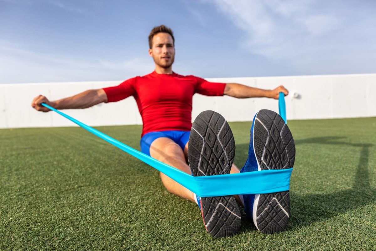 athletic male in red shirt and blue shorts sitting on grass doing lateral arm exercises with resistance band hooked under feet after shoulder replacement surgery