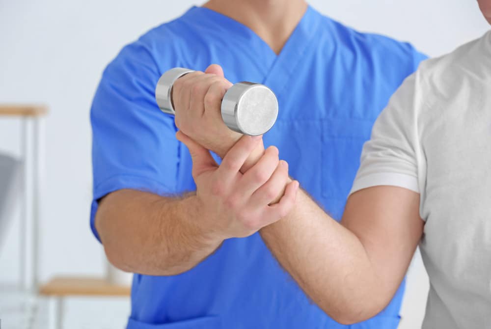 male clinician in blue medical scrubs helping male patient in white shirt complete resistance training during elbow replacement rehabilitation